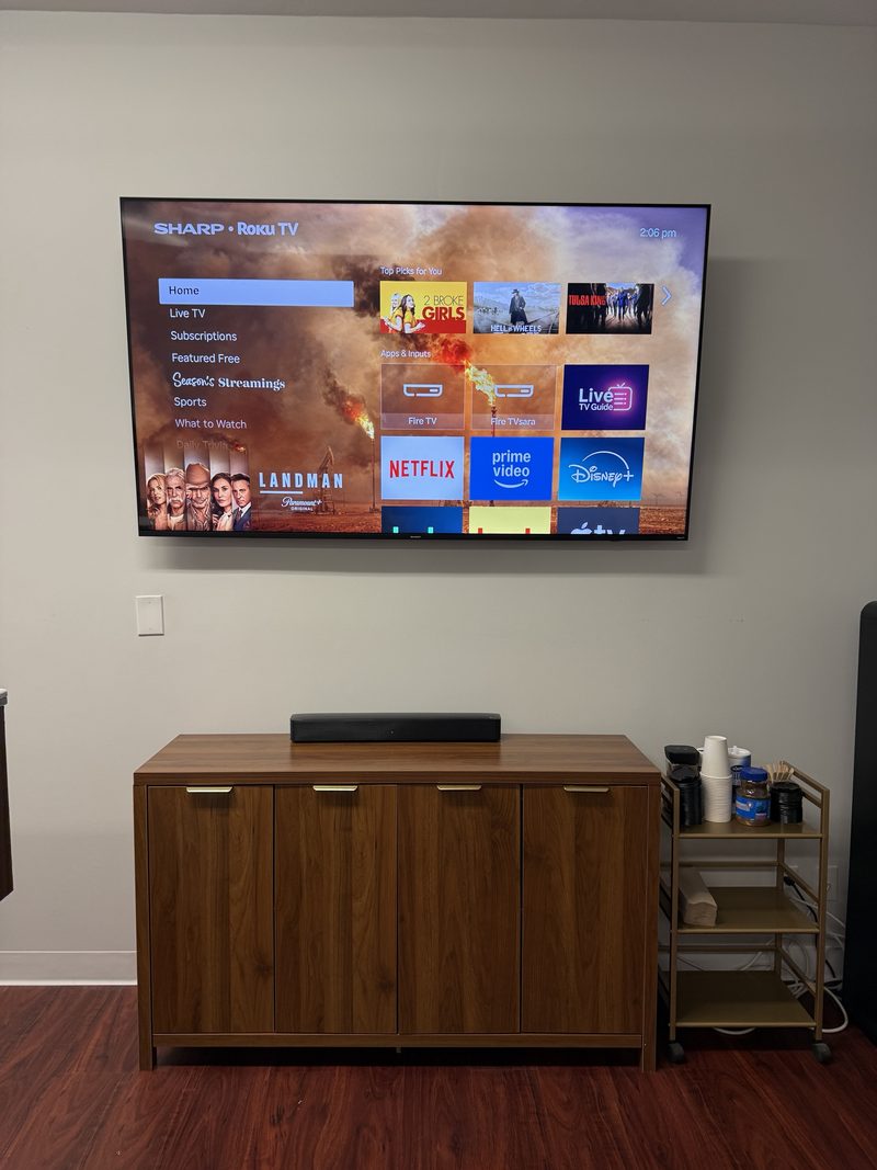 A large flat-screen TV mounted on a wall above a wooden credenza with a soundbar, in a break room.